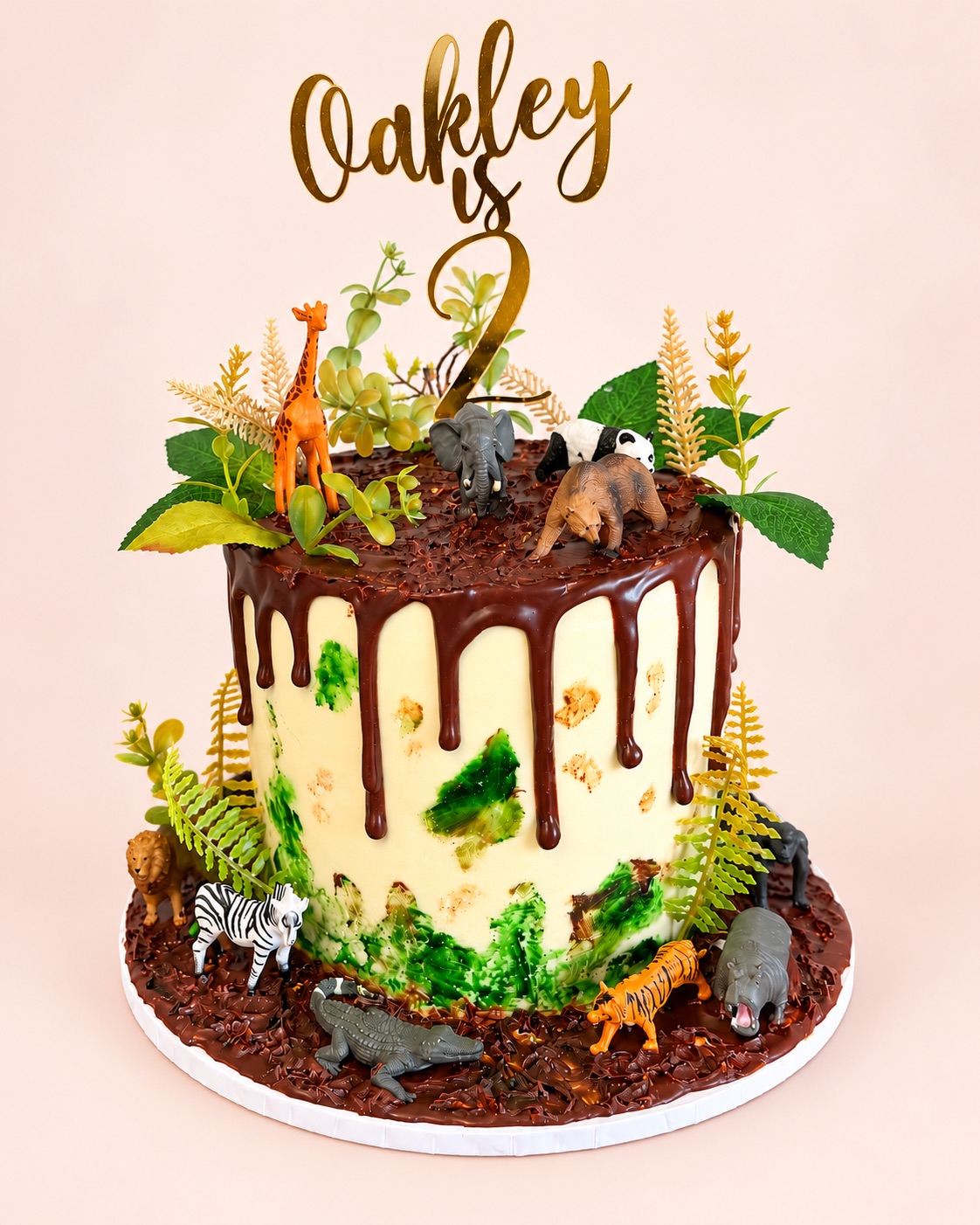 Safari-themed bespoke cake with a chocolate drip, greenery, toy animals and an “Oakley is 2” topper