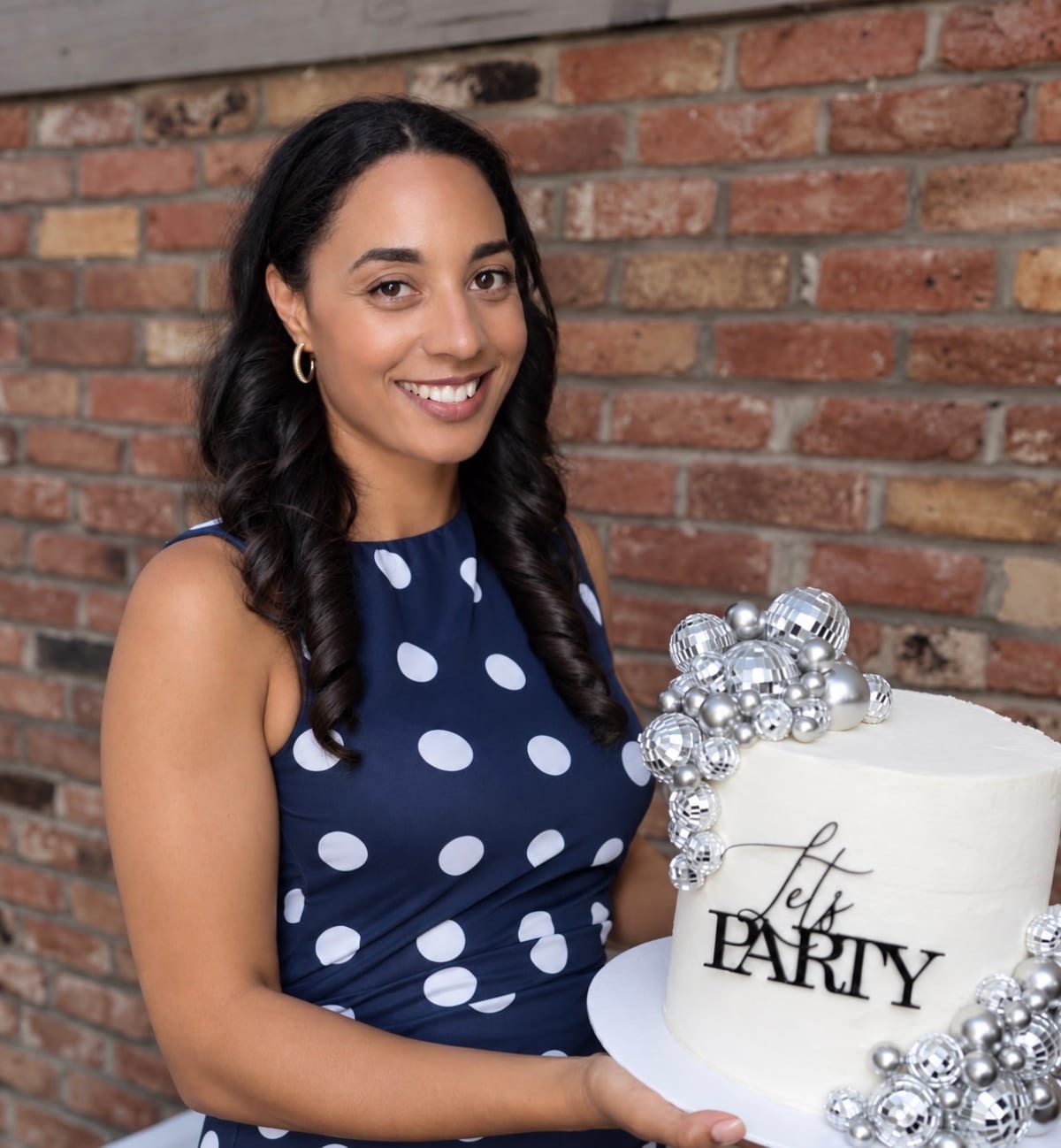 Charlotte smiling and holding a white buttercream cake topped with silver disco balls and a “Let’s Party” topper