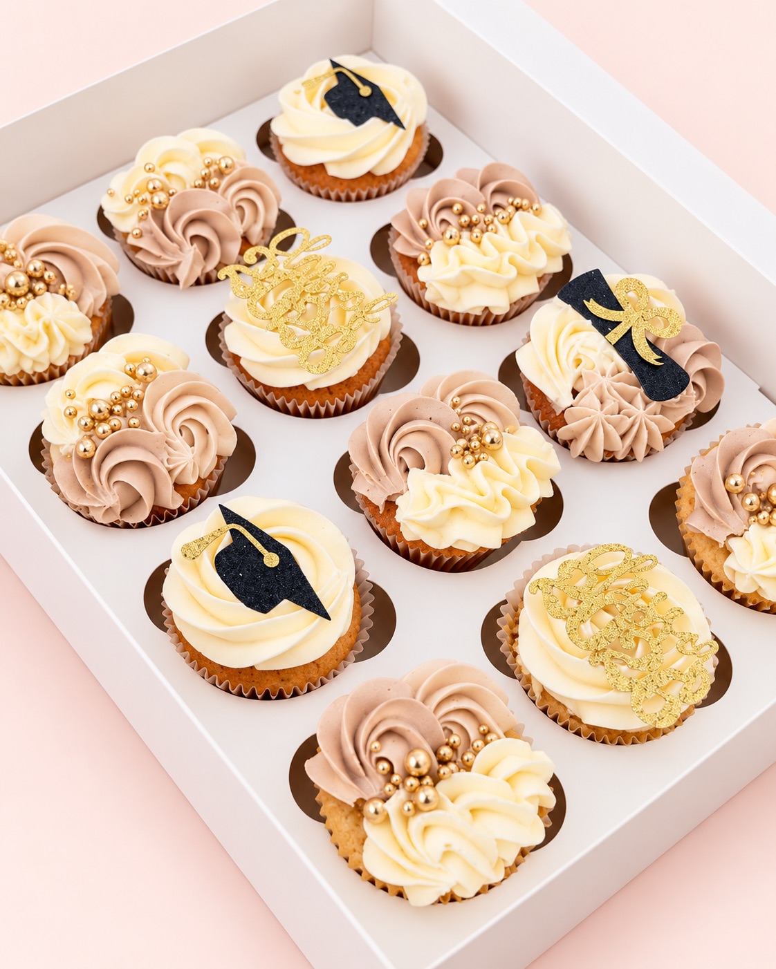 Box of a dozen cupcakes with cream and pink buttercream swirls, gold pearls and graduation cap toppers